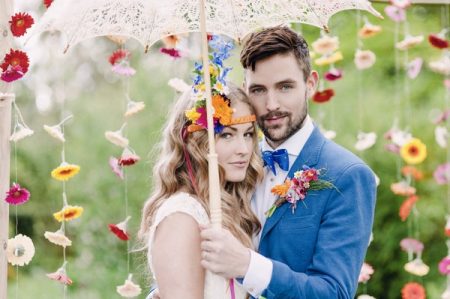 Boho bride and groom under parasol with colourful suspended flower backdrop