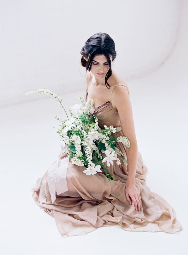 Bride sitting on floor wearing blush dress and holding white and green bouquet