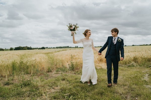 Bride holding bouquet in air as she walks in field with groom