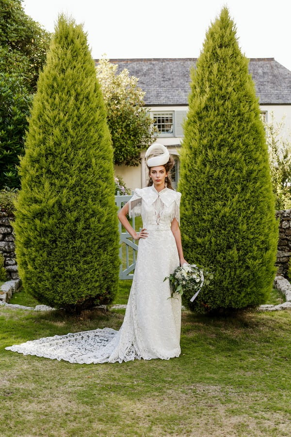 Bride with bridal hat standing in front of two tall bushes at Cosawes Barton