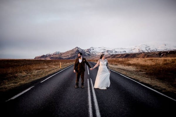 Bride and groom holding hands in the middle of road with mountains in background - Picture by Gavin Jacob Power