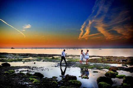 Groom walking across beach towards bride and bridesmaid with beautiful colourful sky - Picture by Marius Tudor Photography