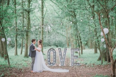 Bride and groom standing by LOVE letters in woodland with bride's dress amking shape of the L - Picture by Ella Parkinson Photography