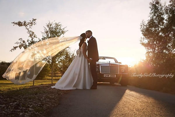 Bride and groom kissing by wedding car with bride's veil blowing in the wind - Picture by Twirly Girl Photography