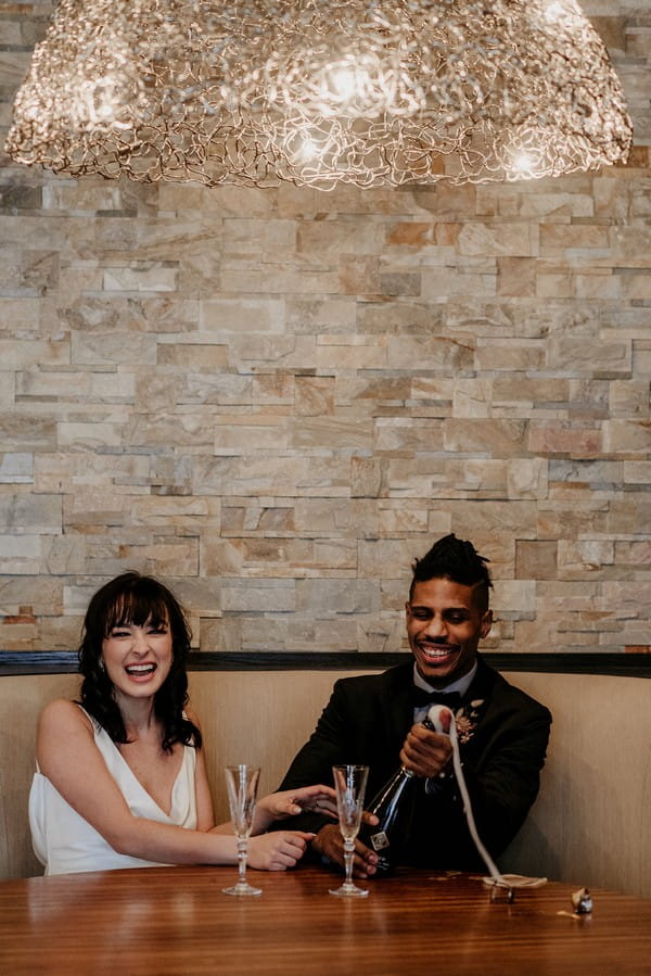 Bride and groom laughing as groom opens bottle of champagne