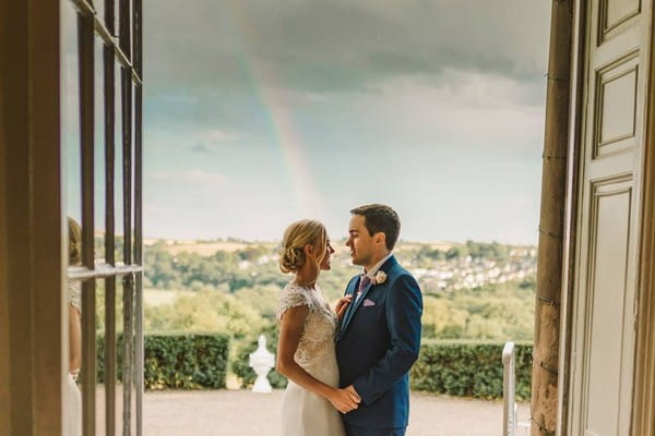 Bride and groom standing in doorway with rainbow in background - Picture by Anna Wood Photography