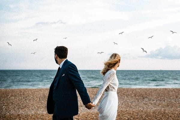 Bride and groom holding hands turning round to see seagulls flying over sea - Picture by Beatrici Photography