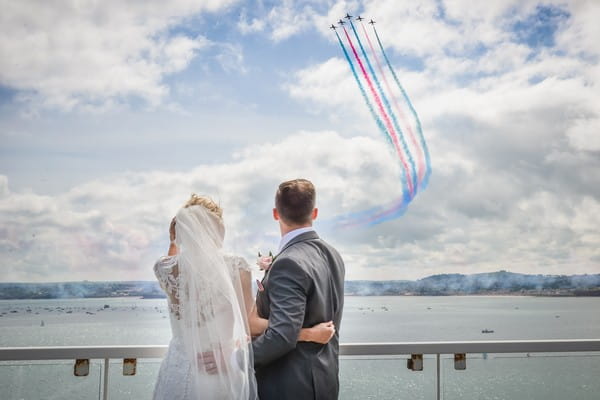 Bride and groom watching as jet planes fly overhead leaving red and blue trails behind them - Picture by Memories and Milestones Photography