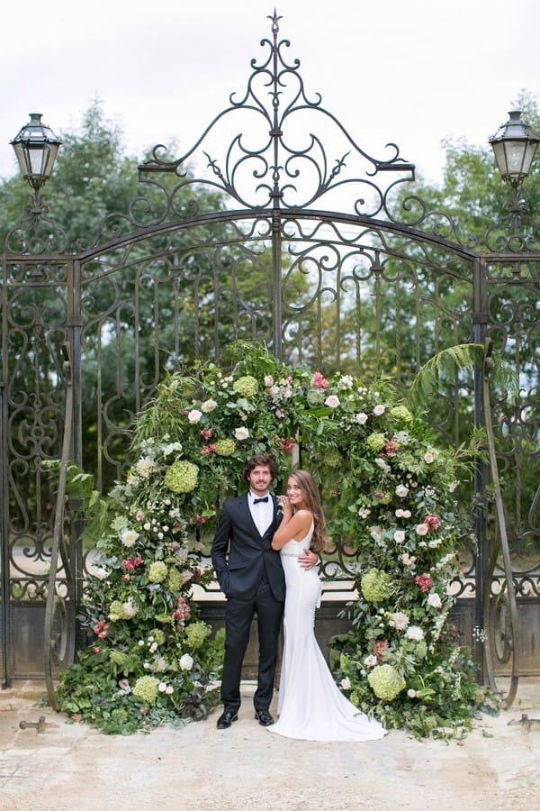 Bride and groom in front of large flower and foliage arch on gates of Chateau de Redon
