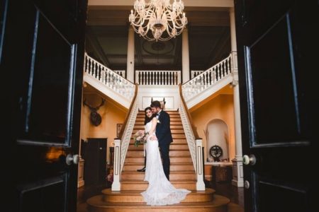 Bride and groom standing at bottom of staircase in entrance of wedding venue - Picture by Gina Manning Photography