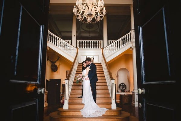 Bride and groom standing at bottom of staircase in entrance of wedding venue - Picture by Gina Manning Photography