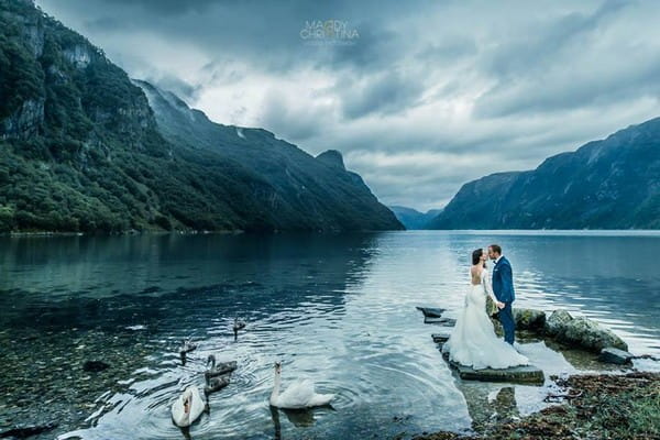 Bride and groom standing by lake surrounded by mountains - Picture by Maddy Christina Photography