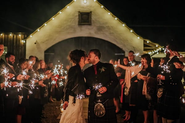 Bride in leather jacket kissing groom in kilt surrounded by guests with sparklers - Picture by Rebecca Rose Noller Photography