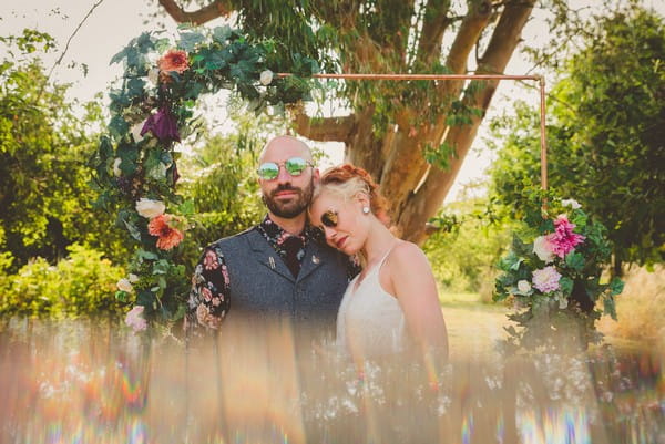 Bride and groom in sunglasses in front of arch made from copper pipes and flowers - Picture by Photography by Paloma