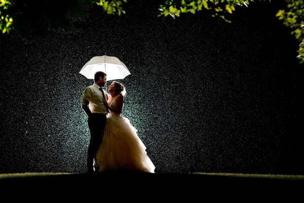 Bride and groom standing under umbrella in the rain at night - Picture by Matt Selby Photography