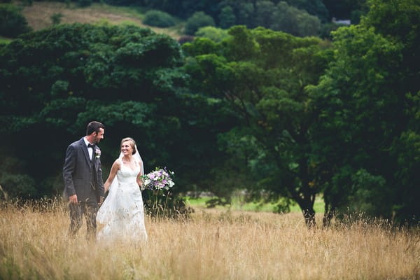Bride and groom holding hands walking through field - Picture by Katie Sidell Photography