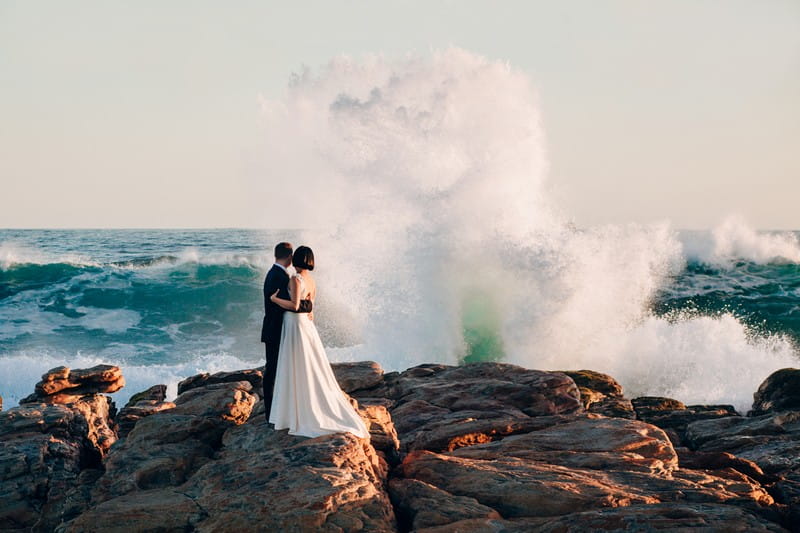 Bride and groom standing on rocks as big wave crashes in front of them - Picture by Estefania Romero Photography