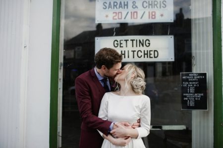 Bride and groom kissing in front of window with Getting Hitched sign - Picture by Jess Petrie Photography