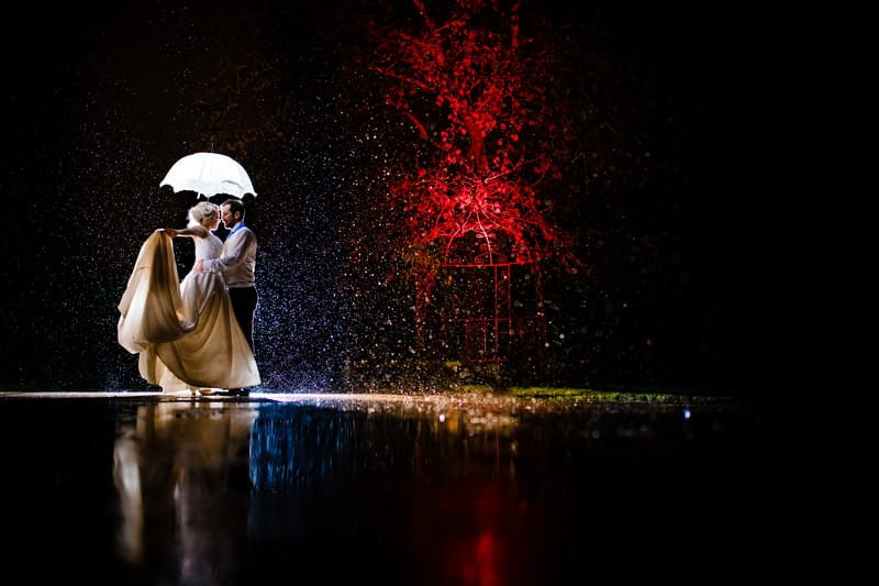 Bride and groom under umbrella in rain at night next to tree lit by red light - Picture by Kimberley Hill Photography