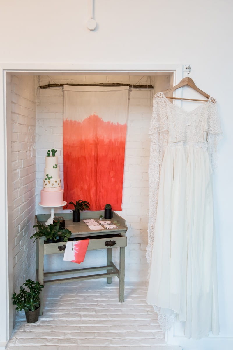 Wedding dress hanging by table with cactus wedding cake and stationery