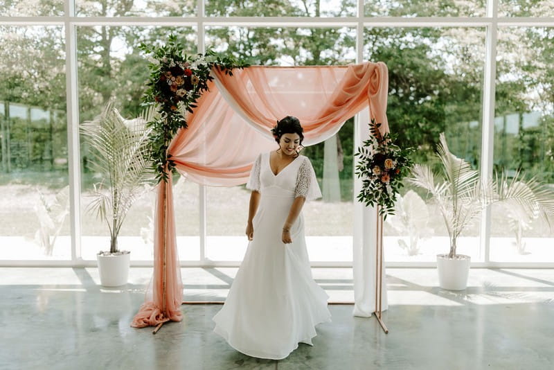 Bride standing in front of ceremony backdrop of pink fabric, foliage and flowers