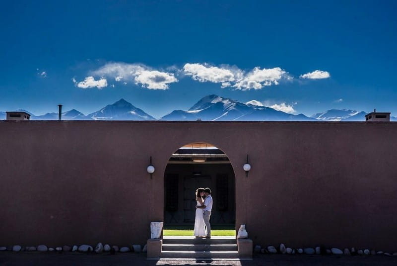 Bride and groom standing in arch opening in wall with mountains in background - Picture by Adrián Zussino Fotografía