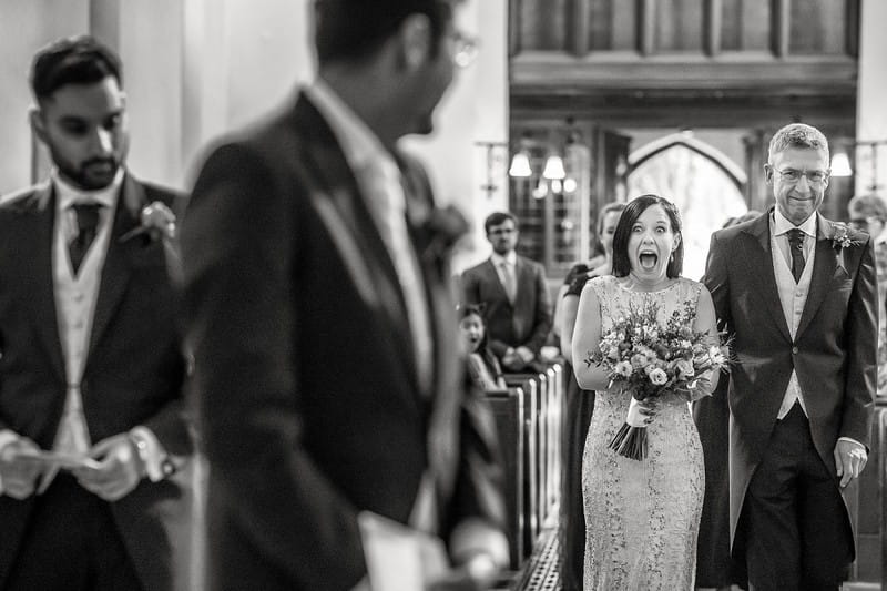 Bride pulling excited face at groom as she walks down the aisle towards him - Picture by Rafe Abrook Photography