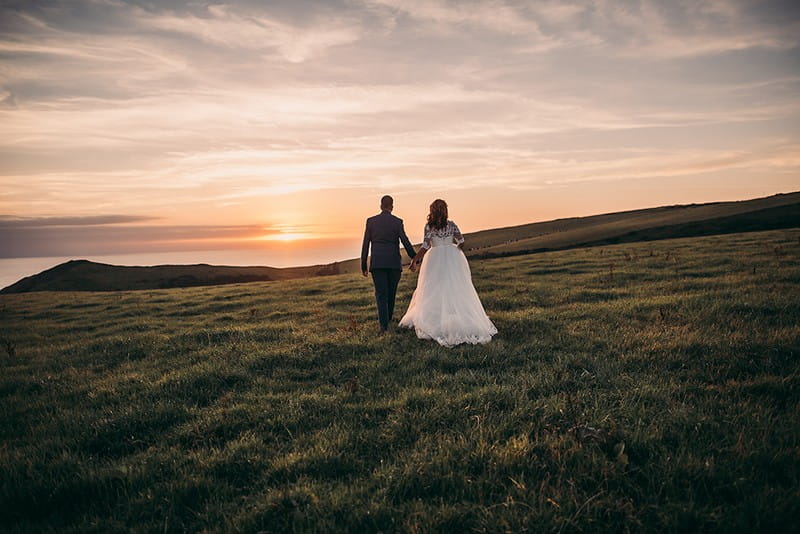 Bride and groom holding hands as they walk across field towards sunset - Picture by Tracey Warbey Photography