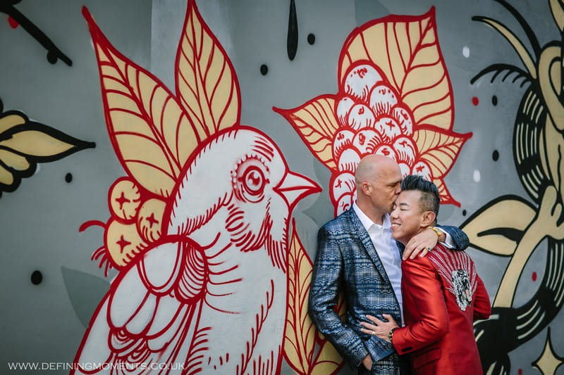Man kissing husband's head against bird graffiti wall - Picture by Defining Moments Photography