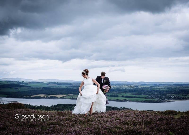 Bride lifting skirt to walk across hill with beautiful view in background - Picture by Giles Atkinson Photography