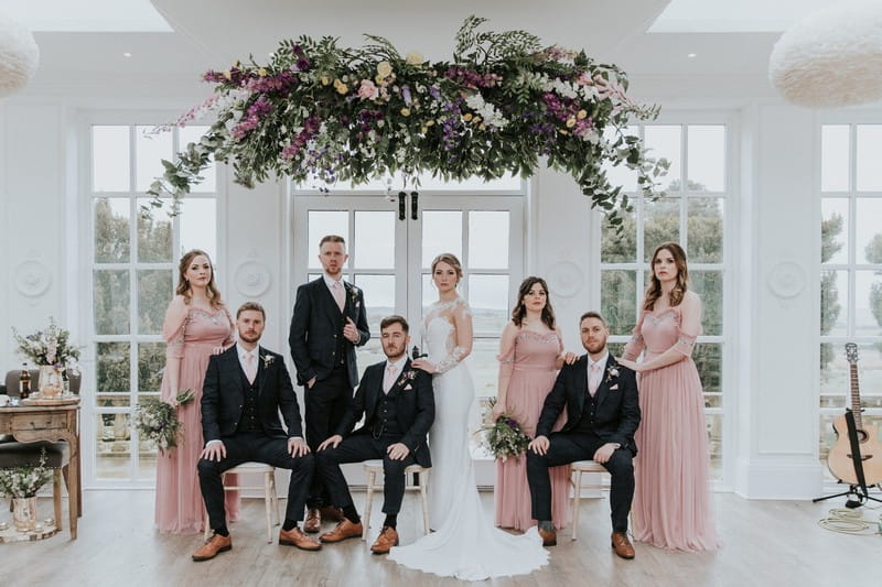 Bridal party posing for photograph under floral chandelier - Picture by Angela Waites Photography