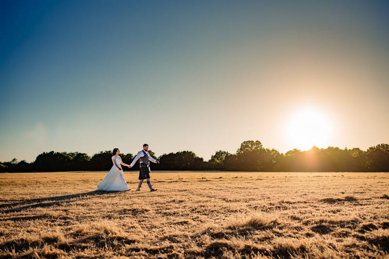 Bride and groom walking across field in hazy sunshine - Picture by Andy Dane Photography