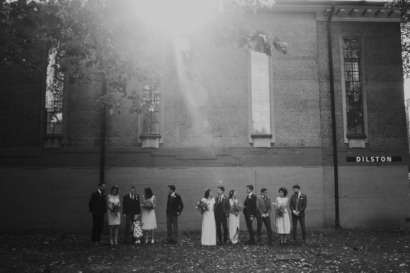 Old style black and white picture of wedding party standing in front of wall of building - Picture by Joasis Photography