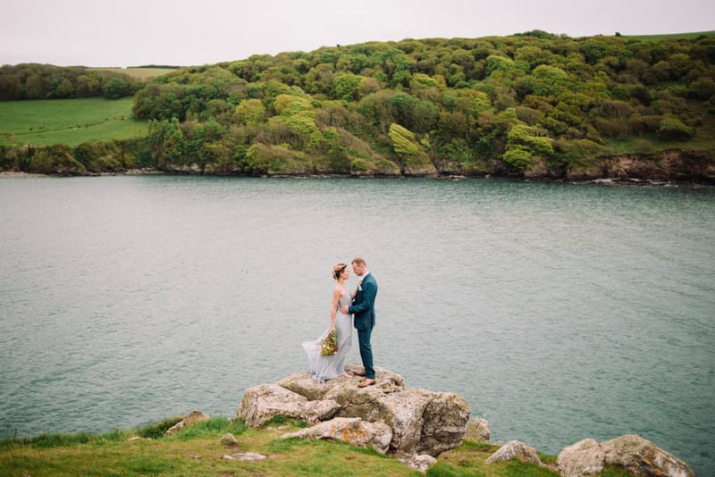 Bride and groom standing on rock by Devon coastline