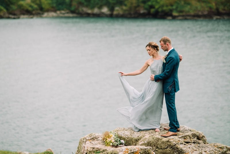 Bride holding out dress as she stands on rock with groom