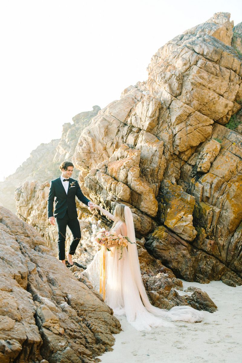 Groom holding bride's hand to help her up onto rocks on beach