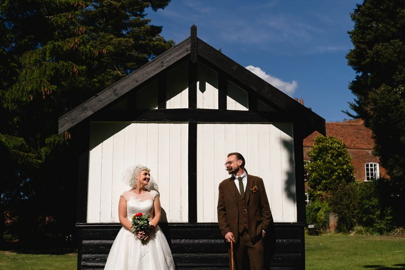 Vintage bride and groom standing up against white wooden building in garden - Picture by Sam Gibson Photography