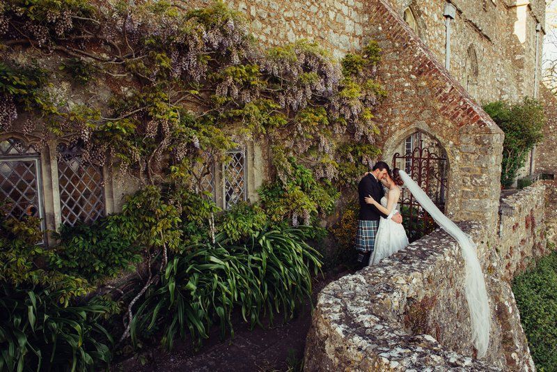 Bride and groom hugging outside venue with bride's veil hanging over wall - Picture by Emma-Jane Photography