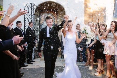 Bride and groom with arms in the air as they walk through confetti shower - Picture by Estefania Romero Photography