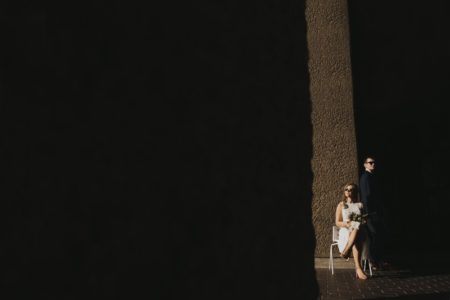 Bride sitting and groom standing in light of dark corridor - Picture by Joasis Photography
