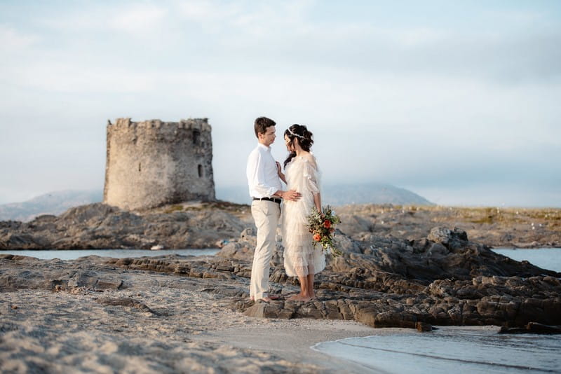 Bride and groom standing on La Pelosa beach with Torre della Pelosa in background