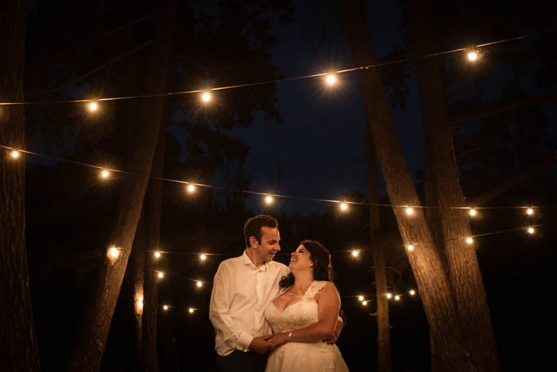 Bride and groom by trees at night under festoon lighting