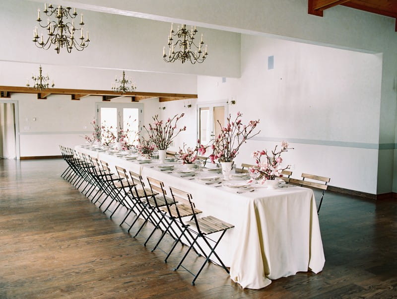 Long wedding table with white tablecloth topped with displays of pink magnolia