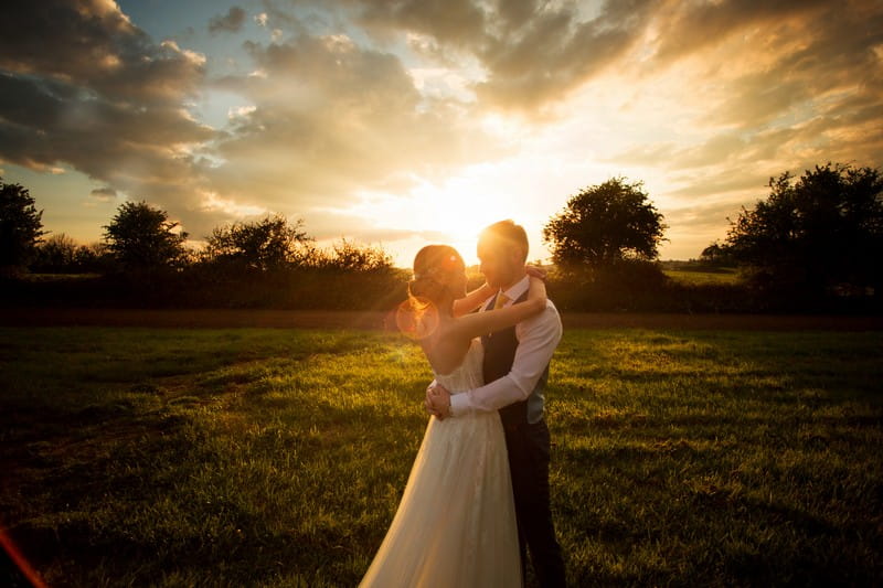 Bride and groom standing in field with arms around each other at sunset - Picture by Martin Dabek Photography