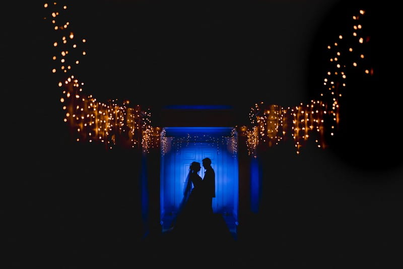 Silhouette of bride and groom against blue light background sourrounded by fairy lights - Picture by Murray Clarke