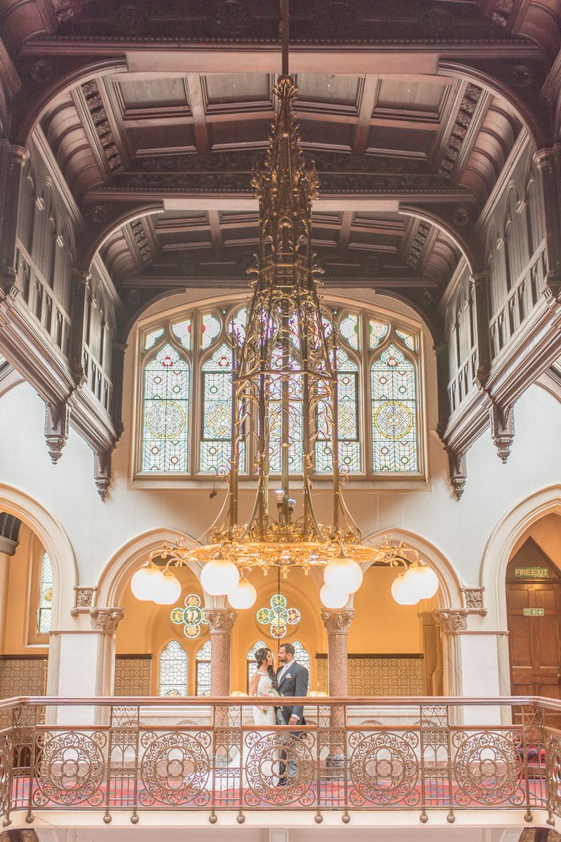 Bride and groom standing under large, elaborate light in grand wedding venue - Picture by Tanya Flannagan Photography