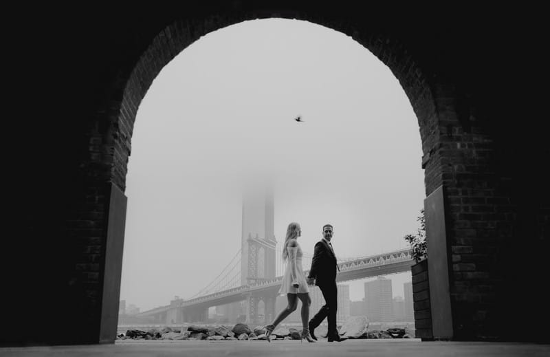 Bride and groom walking past arch in New York with bridge in the background - Picture by Ash Davenport of MIKI Studios