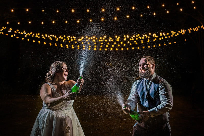Bride and groom spraying champagne in the dark under fairy lights - Picture by Chad Winstead Photography