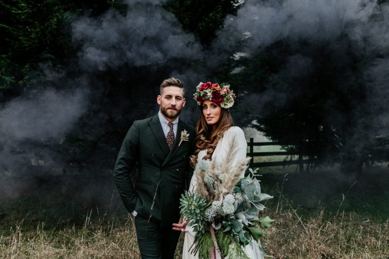 Bride and groom standing in field in front of black smoke - Picture by Epic Love Story