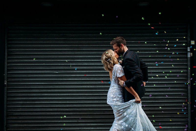 Bride and groom kissing in front of shop shutter with coloured paper falling - Picture by Joanna Bongard Photography